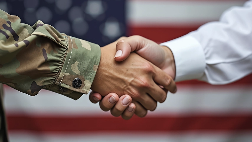 A soldier in camouflage uniform shaking hands with a civilian in a white dress shirt, with an American flag in the background